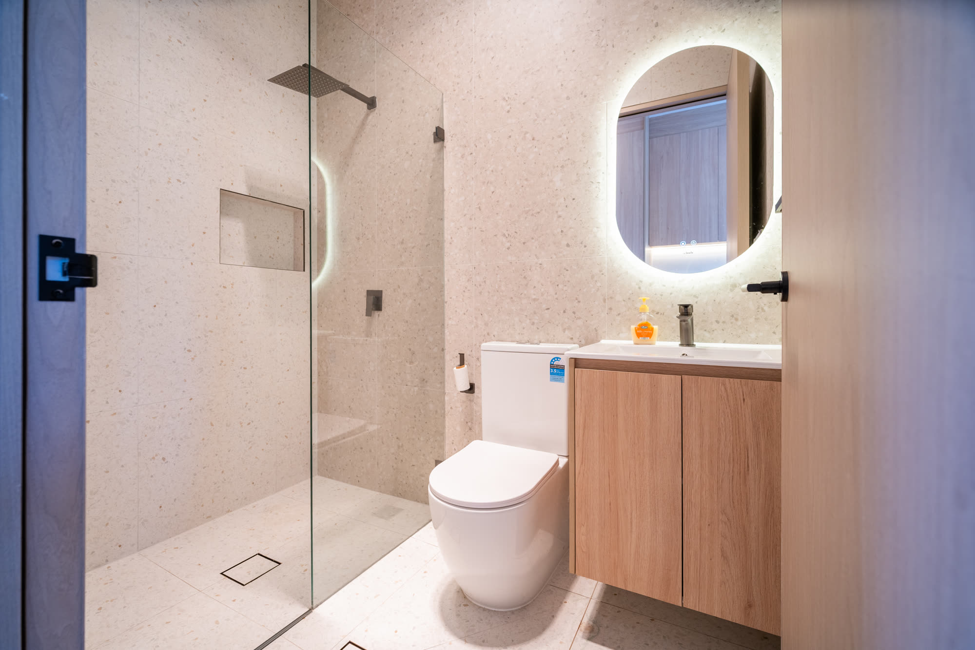 Private ensuite in a completed Auzzy Projects co-living home in Western Sydney, with a walk-in shower, wall-hung timber vanity and a backlit round mirror
